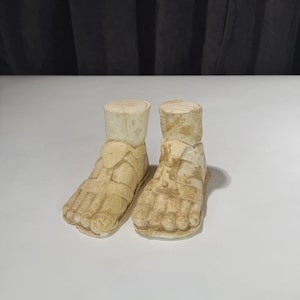 May include: A pair of antique, beige-colored stone sculptures of feet wearing sandals. The sculptures are weathered, with visible texture and detail, and are set against a white background. The sandals have straps and are attached to cylindrical ankle supports.