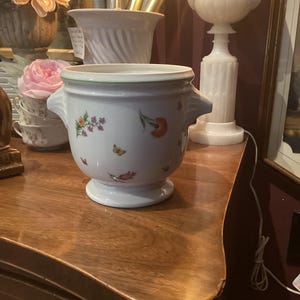 May include: A white ceramic planter with a green rim and two side handles. The planter is decorated with colorful floral and butterfly motifs. It sits on a pedestal base and is displayed on a wooden surface.