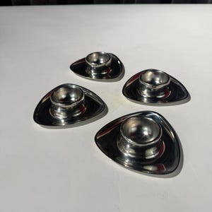 May include: A set of four polished stainless steel egg cups, each featuring a rounded triangular base and a central circular holder. The metallic surface is reflective, showing smooth curves and a consistent, clean design across all four pieces.