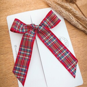 May include: A white postcard with a red and green plaid ribbon tied in a bow. The ribbon is tied in the center of the postcard, with the tails of the ribbon extending to the bottom corners of the postcard. The postcard is on a wooden surface.