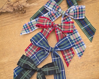 May include: Four plaid fabric bows in green, red, blue, and black, arranged on a wooden surface. Each bow has long, pointed tails. The bows feature a classic tartan pattern, with intersecting stripes of various colors.