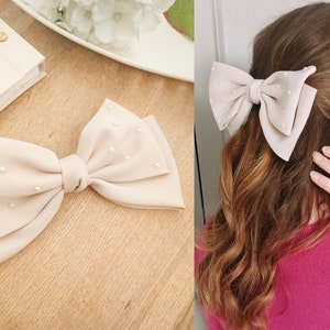 May include: A large beige bow hair clip with pearl embellishments. The bow is made of a soft fabric and has a simple, elegant design.
