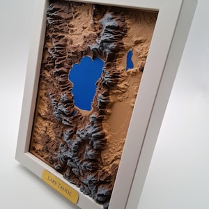 May include: A framed 3D topographical map of Lake Tahoe. The map features a detailed relief of the surrounding mountains in shades of brown and gray, with a vibrant blue lake. The frame is white, and a label at the bottom reads "LAKE TAHOE."