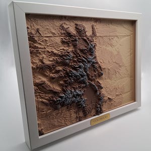 May include: A framed 3D relief map of Colorado, featuring detailed topographical features in shades of brown and blue. The map is set within a white frame, with a gold-colored label that reads "Colorado".
