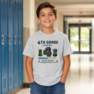 May include: A light gray short-sleeve t-shirt featuring the text 4TH GRADE LOADING... with a large number 4, a pixelated green creature, a block, and a loading bar. Below, the text reads BIGGER DREAMS STRONGER EVERY DAY with two pickaxes.