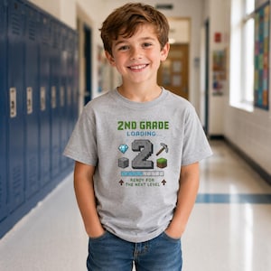 May include: A heather gray short-sleeve t-shirt featuring a graphic with the text 2ND GRADE LOADING... and READY FOR THE NEXT LEVEL. The design includes a large pixelated number 2, a diamond, a pickaxe, and two different block icons.