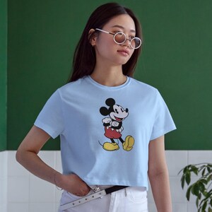 Może przedstawiać: Jasnoniebieski t-shirt z krótkim rękawem z nadrukiem graficznym przedstawiającym Myszkę Miki z przodu. Postać jest ukazana w klasycznym stylu z czarnymi uszami, czerwonym dołem i żółtymi butami, stojąca ze skrzyżowanymi na piersi rękami.