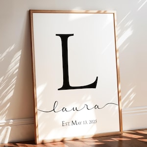 May include: A framed print with a black letter L and the name "laura" written in cursive script. The text "Est. May 13, 2025" is printed below the name.