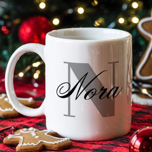 May include: White ceramic mug with the name "Nora" in a script font, with a large gray letter "N" as the initial. The mug is on a red and green plaid surface with gingerbread cookies and Christmas ornaments in the background.