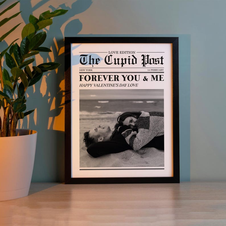 May include: A framed black and white print titled "The Cupid Post" with the text "FOREVER YOU & ME" and "HAPPY VALENTINE'S DAY LOVE." The image shows a couple lying on a beach. A green plant in a white pot is in the background.