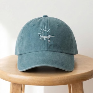 May include: A teal baseball cap with a white line drawing of a sun setting over the ocean. The cap is made of a soft material and has a curved brim. The cap is sitting on a wooden stool.