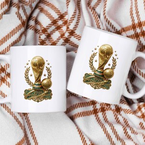 May include: Two white ceramic mugs featuring a gold-colored trophy graphic with a soccer ball, laurel branches, and stars. Below the trophy, a green ornate plaque displays the text 2026 WORLD CUP in gold lettering on both of the mugs.