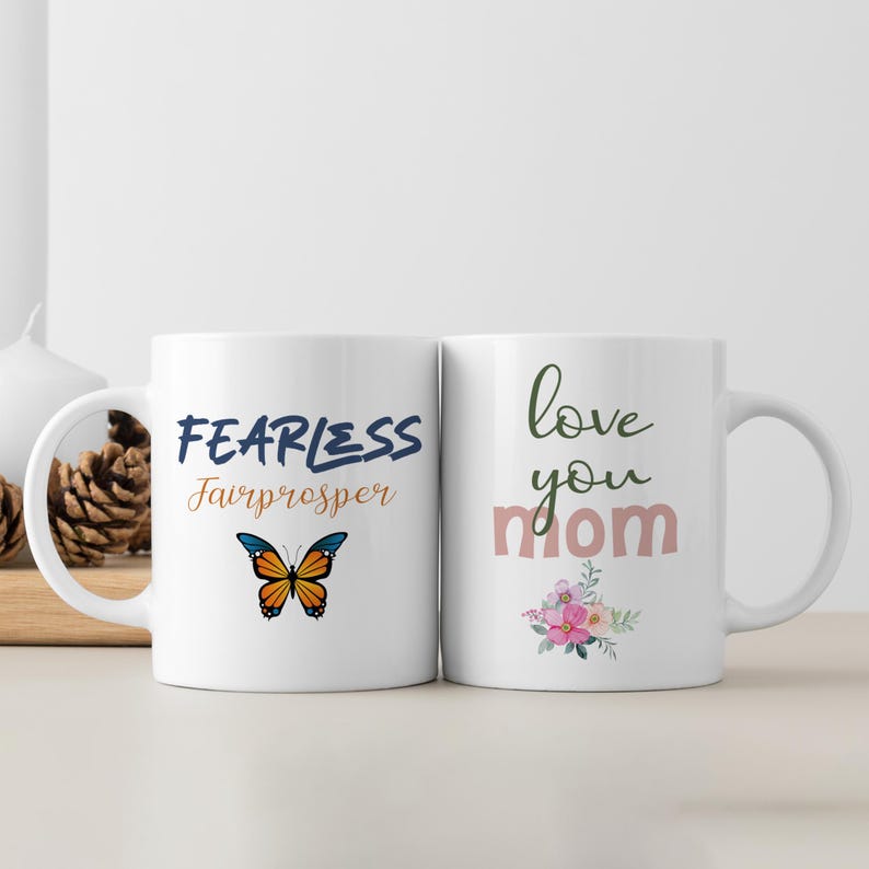May include: Two white ceramic mugs with handles. The mug on the left has a blue and orange butterfly and the text "FEARLESS Fairprospter". The mug on the right has pink flowers and the text "love you mom".
