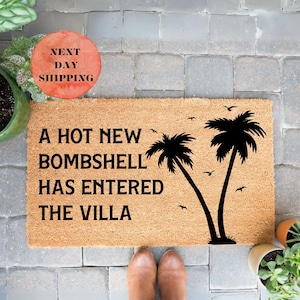 May include: A rectangular tan coir doormat featuring the black text A HOT NEW BOMBSHELL HAS ENTERED THE VILLA, accompanied by a black graphic of two palm trees and several small birds flying in the sky on the right side of the mat.