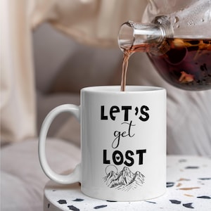 May include: White ceramic coffee mug with a black design that says "Let's get lost" with a mountain range and a heart.