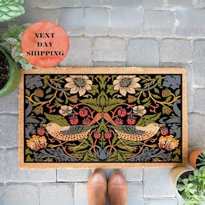 May include: A coir doormat with a black background and a colorful floral and bird design. The design features two birds, flowers, and berries in shades of red, yellow, green, and blue.
