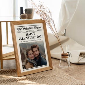 May include: A framed print of "The Valentine Times" newspaper with the headline "Happy Valentine's Day!" and a photo of a couple. The frame is made of light-colored wood. A glass vase with dried stems is to the right.