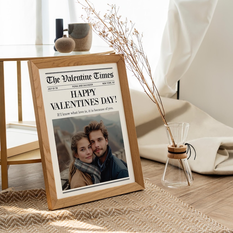 May include: A framed print of "The Valentine Times" newspaper with the headline "Happy Valentine's Day!" and a photo of a couple. The frame is made of light-colored wood. A glass vase with dried stems is to the right.