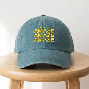 May include: A teal baseball cap with the word "AMAZE" embroidered in yellow. The word is repeated three times, with a curved line around the text. The cap is sitting on a wooden stool.