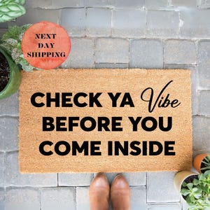 May include: A rectangular doormat made of natural fibers with the text "CHECK YA VIBE BEFORE YOU COME INSIDE" in black script. The mat is on a gray brick surface, with plants in pots on either side.