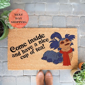 May include: A brown coir doormat with the text "Come inside and have a nice cup of tea!" in black lettering. A blue and purple cartoon worm with a red scarf is on the right side of the doormat.