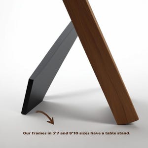 May include: A black and brown wooden picture frame with a table stand. The frame is angled, with a black front and a brown wooden support. Text at the bottom reads, "Our frames in 5*7 and 8*10 sizes have a table stand."