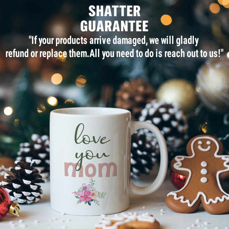 May include: White ceramic mug with a green "love you mom" design and pink floral accents. The mug is sitting on a table with a gingerbread man cookie and pine cones.