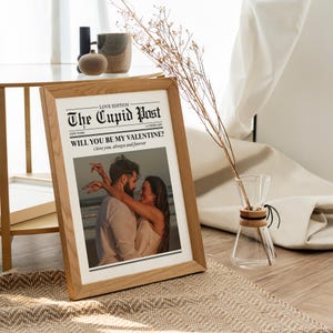 May include: A framed print with a wooden frame. The print is a newspaper-style design titled "The Cupid Post" with the headline "Will You Be My Valentine?" and a photo of a couple embracing. A glass vase with dried stems is next to the frame.