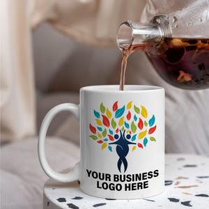 May include: White ceramic mug with a colorful tree design and the text "YOUR BUSINESS LOGO HERE". Tea is being poured into the mug.