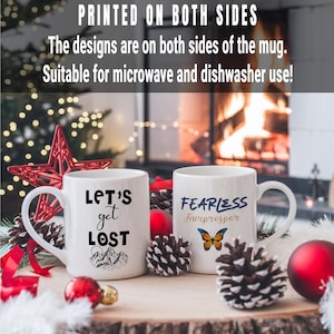 May include: Two white ceramic mugs with black text and designs. The mug on the left says "Let's get lost" with a mountain range design. The mug on the right says "Fearless Fairprosper" with a butterfly design.