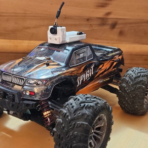 Könnte beinhalten: Ein schwarzer und orangefarbener Monstertruck mit einer auf dem Dach montierten Kamera. Der Truck hat das Wort "SPIRIT" auf der Seite und "4WD RACING" darunter gedruckt.