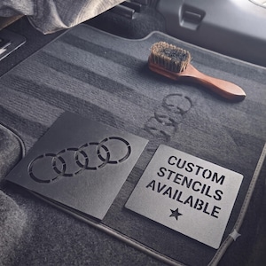 May include: Two square black metal stencils resting on a dark grey car floor mat. One stencil features the four-ring Audi logo, while the other displays the text "CUSTOM STENCILS AVAILABLE" with a star. A wooden brush with dark bristles sits nearby.
