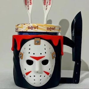 May include: A black mug with a white hockey mask design, red accents, and a handle shaped like a knife. The lid has the word "new!" printed on it. Two white spoons are inside the mug.