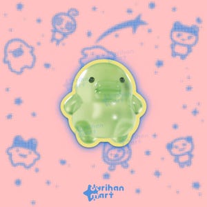 May include: A glossy, light green, rounded duck-shaped figurine with a smooth surface, small black eyes, and a wide beak. The figure is outlined in a thin yellow border and set against a pink background featuring blue dotted patterns and the text durihan mart.
