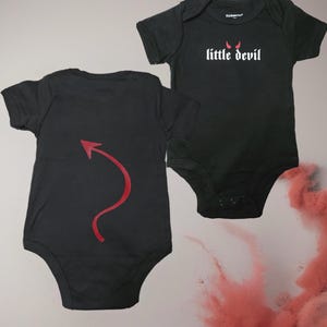 May include: Black baby bodysuit with the text "little devil" in white and red devil horns. The back of the bodysuit has a red arrow-shaped tail design. The bodysuit is made of soft material.