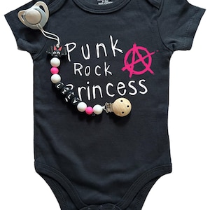 Puede incluir: Body de bebé negro con el texto "Punk Rock Princess" y un símbolo de anarquía rosa. Un chupete y un clip de chupete con cuentas y un dije de murciélago están adjuntos.