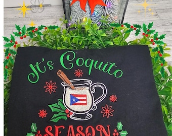 Puede incluir: Sudadera negra con texto y gráficos bordados en verde y rojo. El texto dice "It's Coquito Season" con un diseño de taza con la bandera de Puerto Rico, una rama de canela y acentos de acebo. Ropa festiva de vacaciones.