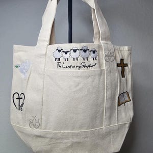 May include: A cream-colored canvas tote bag featuring embroidered designs including a row of five sheep, the text "The Lord is my Shepherd," a white dove with an olive branch, a black heart with the word "Jesus," a brown cross, and an open book.