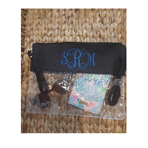 May include: A clear plastic bag with a black zippered top and a black strap. The bag has a black fabric flap with the monogram "SRM" embroidered in blue thread. The bag is filled with sunglasses and a patterned wallet.