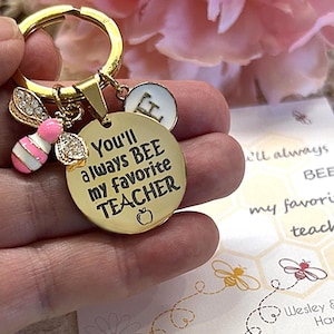 May include: A gold-toned metal keychain featuring a round disc engraved with the text You'll always BEE my favorite TEACHER and a small apple icon. It includes a pink and white enamel bee charm with clear crystals and a small white circular charm.