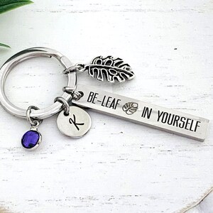 May include: Silver keychain with a charm that says "Be-Leaf in Yourself" and a small leaf charm. There is also a small purple gem and a silver charm with the letter K.
