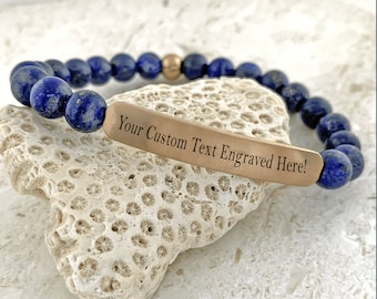 May include: A beaded bracelet featuring dark blue lapis lazuli stones and a gold-colored accent bead. The bracelet includes a rectangular, gold-toned metal plate with the engraved text "Your Custom Text Engraved Here!"