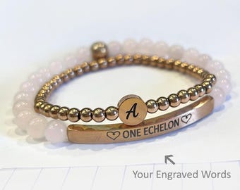 May include: Two bracelets. One is a beaded bracelet with light pink beads. The other is a gold-colored bracelet with the letter "A" on a circular charm and the words "ONE ECHELON" engraved on it. The bracelets are on a white surface.