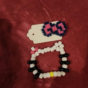 Op de afbeelding: Een kralenarmband met witte, zwarte, roze en gele plastic kralen in een cirkelvormig patroon, vergezeld door een haarspeld gemaakt van donkerblauwe en roze strijkkralen bevestigd op een witte, geschulpte presentatiekaart.