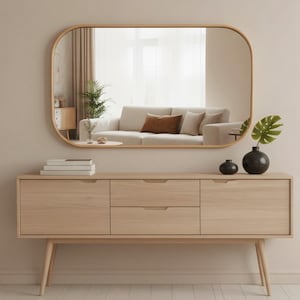 May include: A light wood sideboard with three drawers and two doors, topped with a rounded rectangular mirror. The mirror reflects a living room with a sofa. A black vase and small decorative items sit on the sideboard.