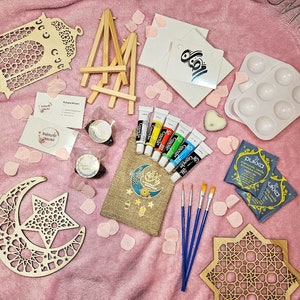 Puede incluir: Un kit de manualidades con un caballete de madera, dos recortes de madera de una luna creciente y una estrella, un lienzo, tubos de pintura, pinceles y una pequeña bolsa de té.