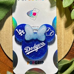 May include: A blue and white phone grip with a glitter finish. The grip features a white bow, a baseball with a heart, and the text "Dodgers" and "LA". The grip is a PopGrip brand.