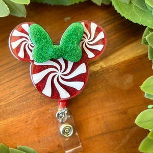 May include: Red and white peppermint candy cane shaped badge holder with a green glitter bow. The badge holder is attached to a clear retractable badge reel.
