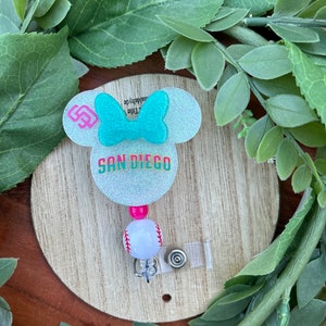 May include: A light blue glitter Minnie Mouse-shaped badge holder with a pink bow, the text "SAN DIEGO" and a baseball charm. The holder has a clear plastic retractable badge clip.