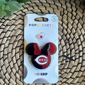 May include: A red and black phone grip with a red glitter finish and a black and red Cincinnati Reds logo. The grip is shaped like Mickey Mouse ears. The packaging says "PopSockets" and "PopGrip".